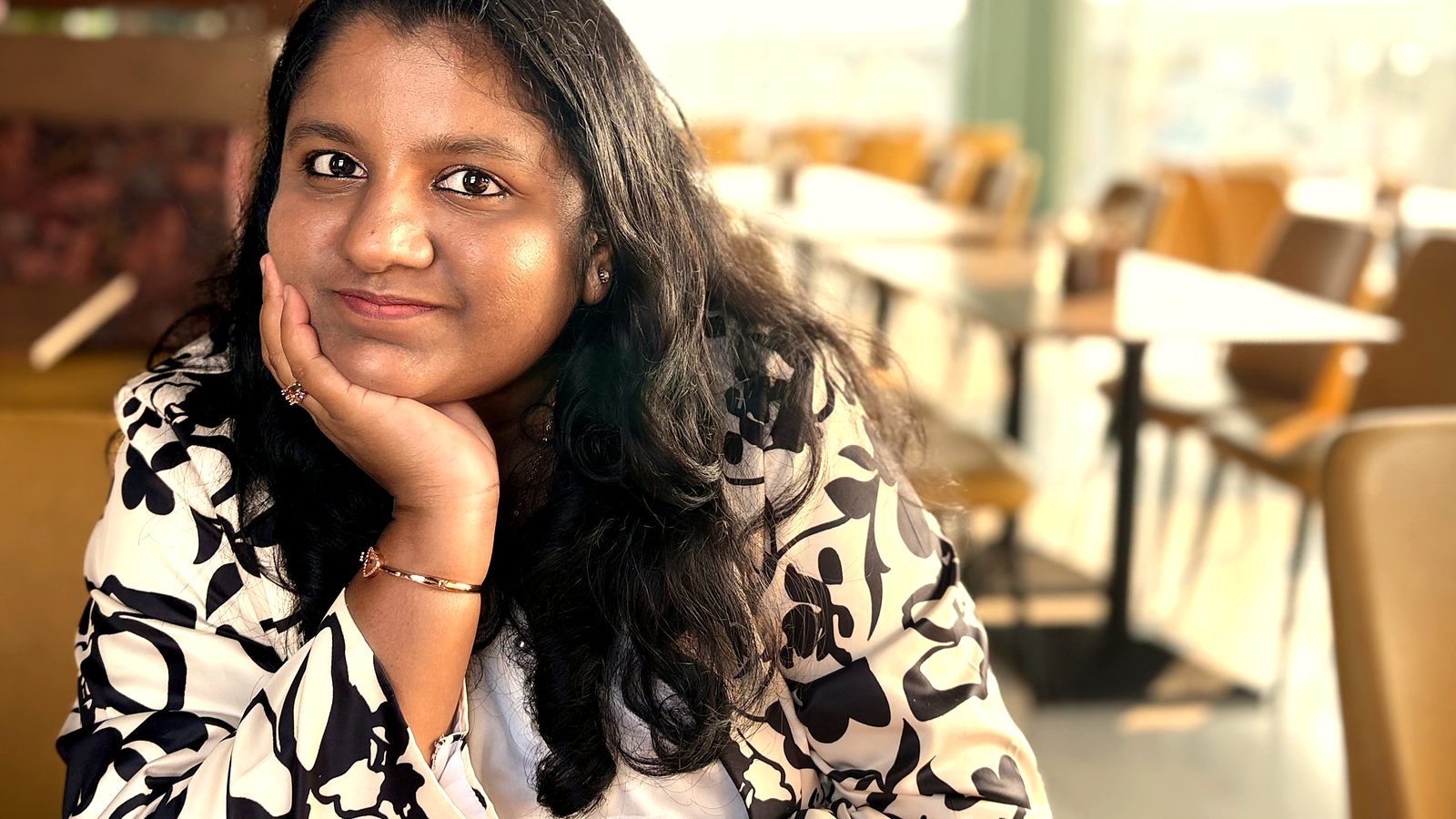 Arthi Rajendran smiling warmly at the camera, chin resting on her hand in a bright cafe