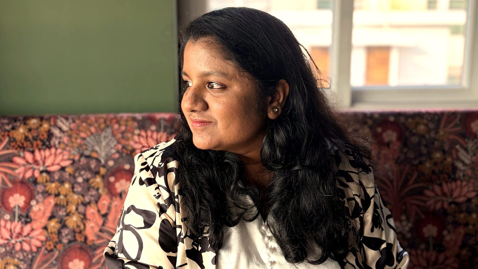 Arthi Rajendran sitting by a bright window, looking away thoughtfully with a calm smile
